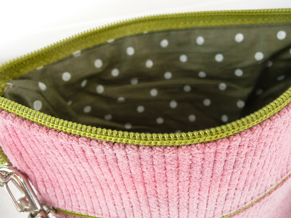 Close-up of a pink ribbed corduroy pouch or bag shown open, with an olive green zipper, white polka dot lining, and a silver metal clasp visible on the left.