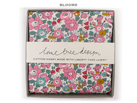 A folded cotton hanky with a colorful floral print in pink, teal, red, and yellow on a cream background, presented in a gray gift box with a white paper band reading 'lime tree design' and 'COTTON HANKY MADE WITH LIBERTY TANA LAWN™'. The word 'BLOOMS' appears above the box.