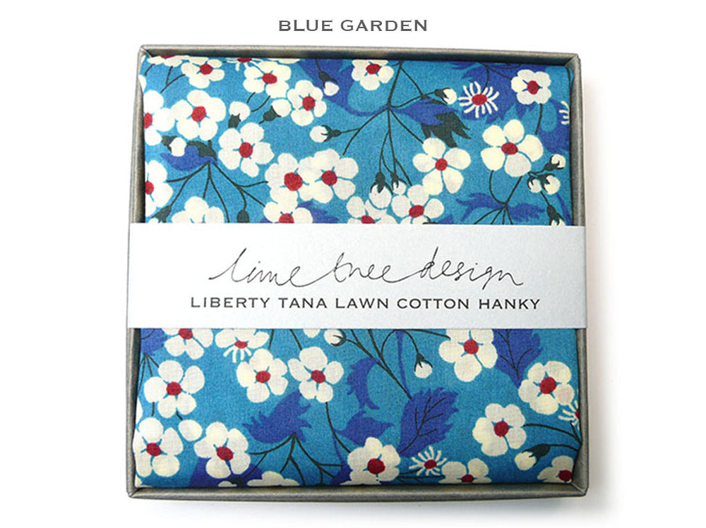 A folded cotton hanky with a blue floral print, presented in a square gray box with a white paper band reading 'lime tree design' and 'LIBERTY TANA LAWN COTTON HANKY.' The top of the image has the text 'BLUE GARDEN.