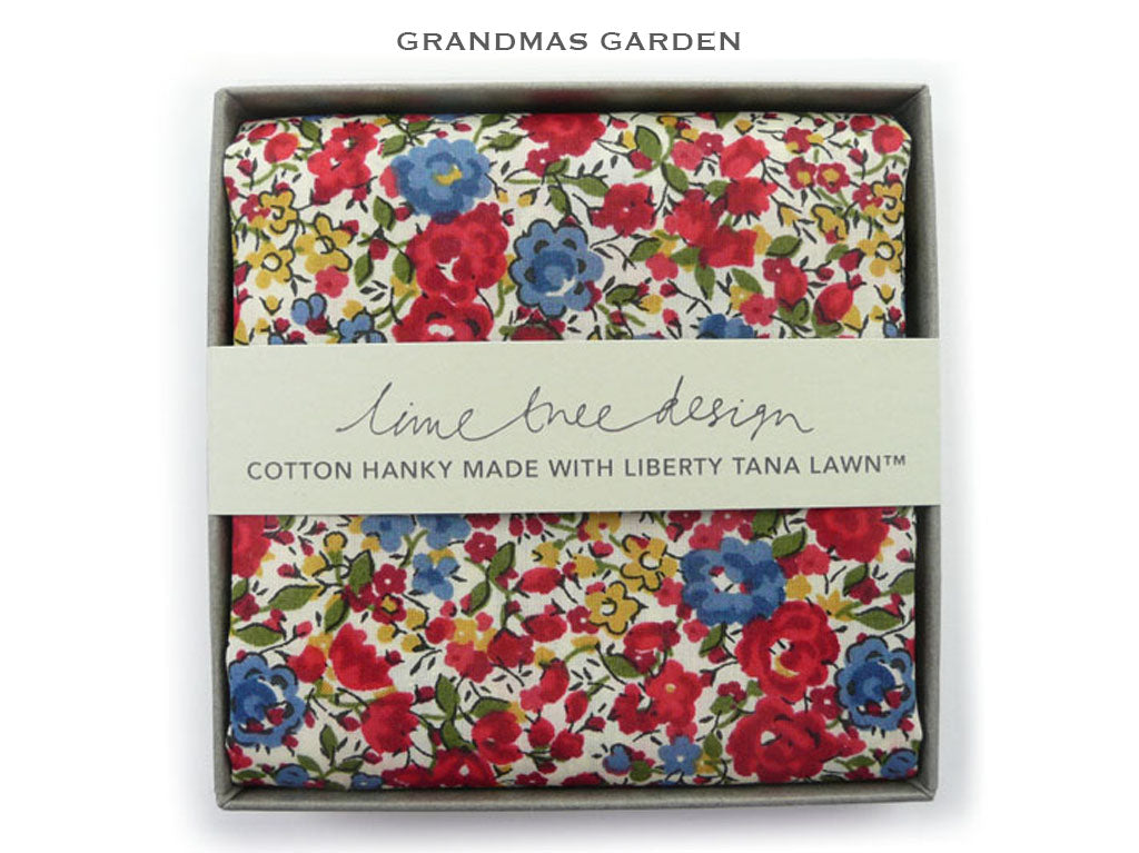 A folded cotton hanky with a vibrant floral print in red, blue, yellow, and green on a white background, presented in a gray box with a paper band reading &