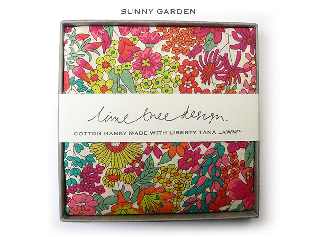 A boxed cotton hanky from Sunny Garden, featuring a vibrant floral pattern in pink, red, orange, green, and yellow on a cream background. The hanky is wrapped in a white paper band labeled &