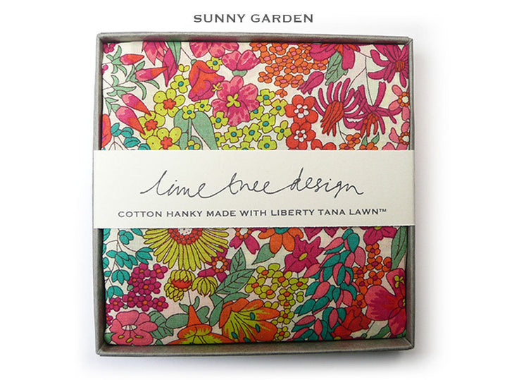 A boxed cotton hanky from Sunny Garden, featuring a vibrant floral pattern in pink, red, orange, green, and yellow on a cream background. The hanky is wrapped in a white paper band labeled &