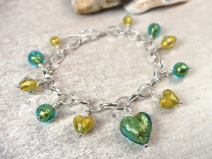 A silver chain bracelet with dangling heart and bead charms in shimmering green, teal blue, and yellow-gold, displayed on a light textured surface.