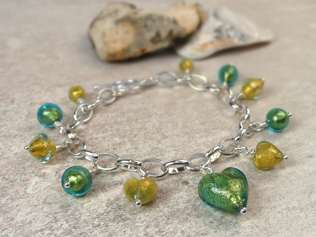 A silver chain bracelet with dangling green and yellow glass beads and a large green heart charm, displayed on a textured stone surface with small rocks in the background.