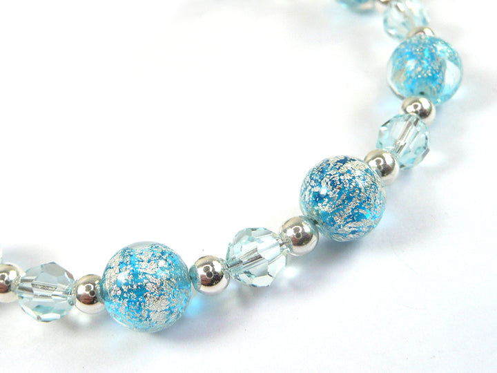 A close-up of a beaded strand featuring light blue glass beads with metallic silver crackle patterns, alternating with faceted clear glass beads and polished silver spacer beads, set against a plain white background.