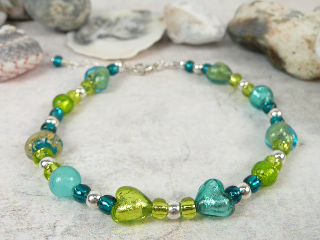 A handmade beaded bracelet with turquoise, green, and silver beads, featuring heart-shaped glass beads, displayed on a light stone surface with seashells in the background.