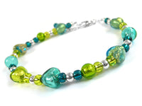 A beaded bracelet featuring translucent green and teal glass beads, including heart-shaped and textured round beads, separated by small metallic silver spacer beads, with a silver clasp at the back, shown on a white background.