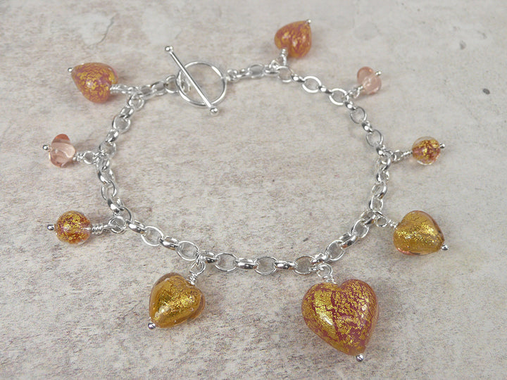 A silver chain bracelet with a toggle clasp and dangling heart-shaped glass charms in amber, gold, and pink tones, displayed on a light textured surface.