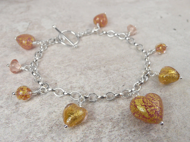 A silver chain bracelet with toggle clasp, decorated with multiple dangling heart-shaped and round beads in metallic gold and orange with a glassy texture, displayed on a light textured surface.