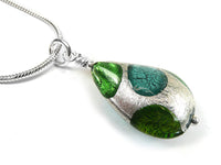 A close-up of a teardrop-shaped glass pendant necklace with green and teal glass inclusions and a textured silver-tone metal base, hanging from a silver chain on a white background.