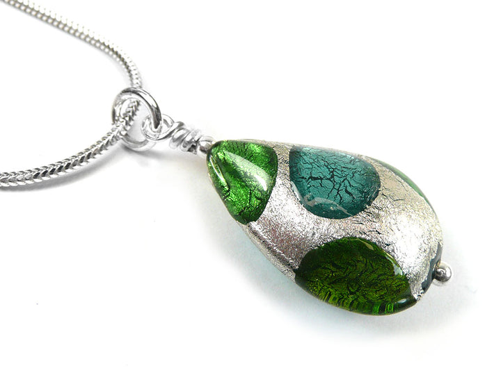 A close-up of a teardrop-shaped glass pendant necklace with green and teal glass inclusions and a textured silver-tone metal base, hanging from a silver chain on a white background.