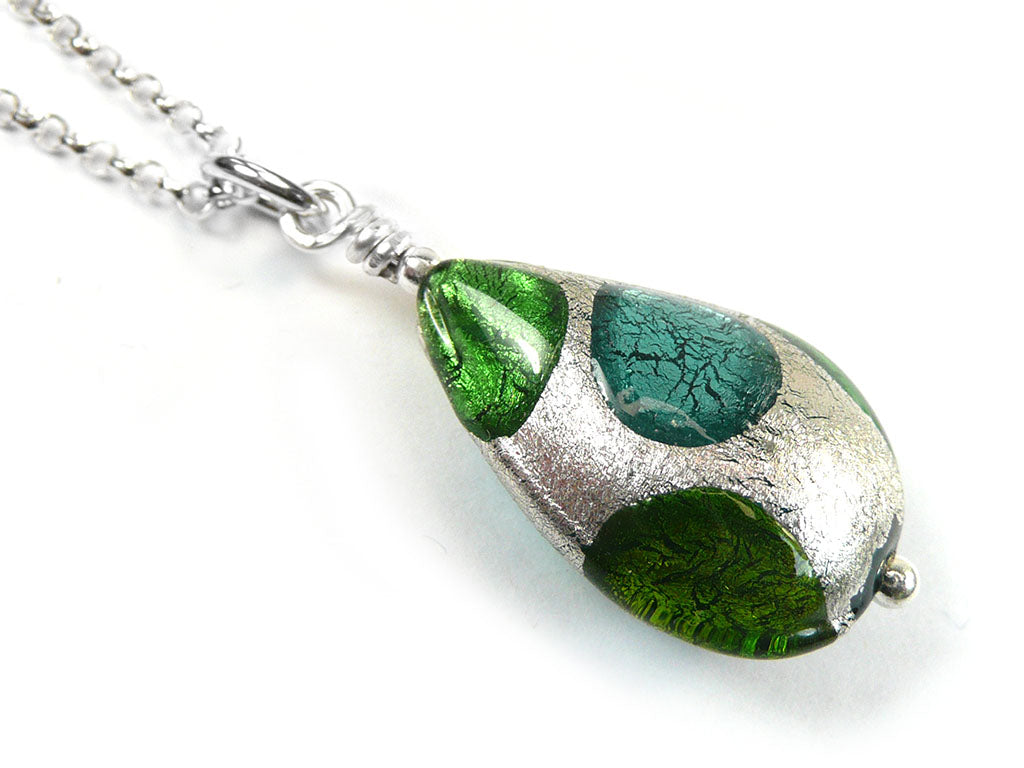 A close-up of a teardrop pendant necklace with a textured silver wrap and three green glass inlays of different shades and patterns, suspended from a silver chain on a white background.