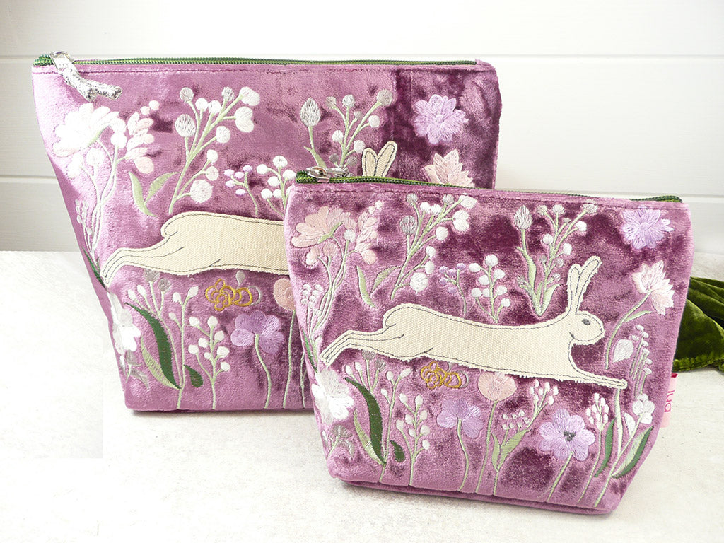Two magenta velvet zippered pouches with embroidered white rabbits, pink flowers, and green leaves, displayed on a white surface against a white tiled wall.