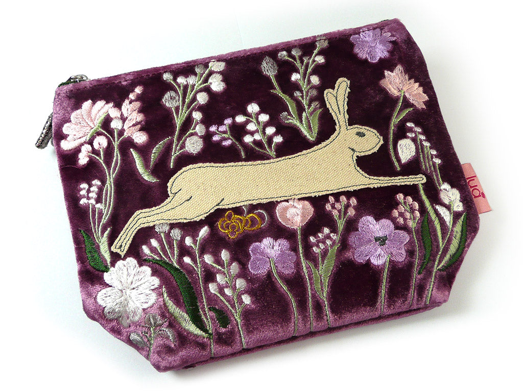 A purple velvet zipped pouch featuring an embroidered design of a beige hare surrounded by pink, white, and purple flowers with green stems, shown on a white background.