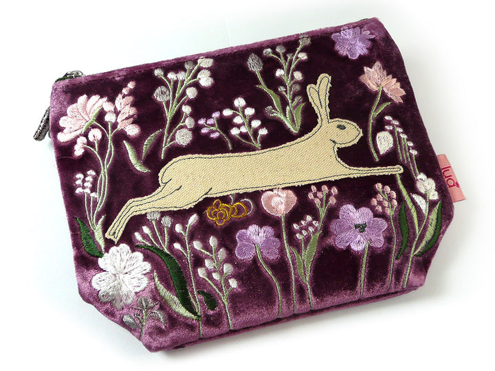 A purple velvet zipped pouch featuring an embroidered design of a beige hare surrounded by pink, white, and purple flowers with green stems, shown on a white background.
