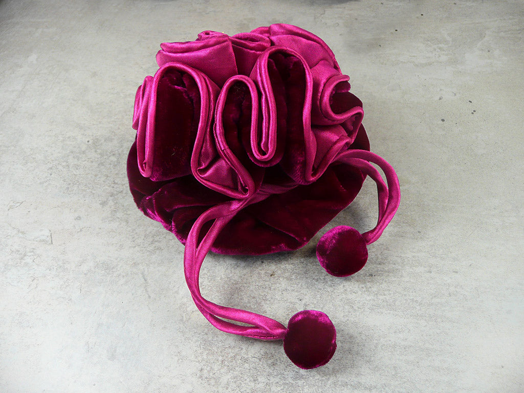 A plush velvet drawstring pouch in a rich magenta pink color, featuring a gathered ruffled design and two matching pom-pom tassels, resting on a textured gray concrete surface.