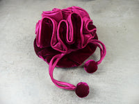 A plush velvet drawstring pouch in a rich magenta pink color, featuring a gathered ruffled design and two matching pom-pom tassels, resting on a textured gray concrete surface.