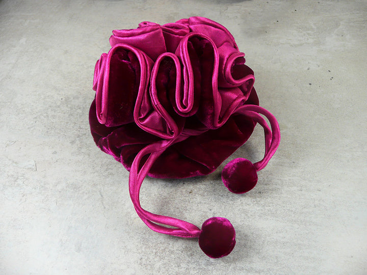 A plush velvet drawstring pouch in a rich magenta pink color, featuring a gathered ruffled design and two matching pom-pom tassels, resting on a textured gray concrete surface.
