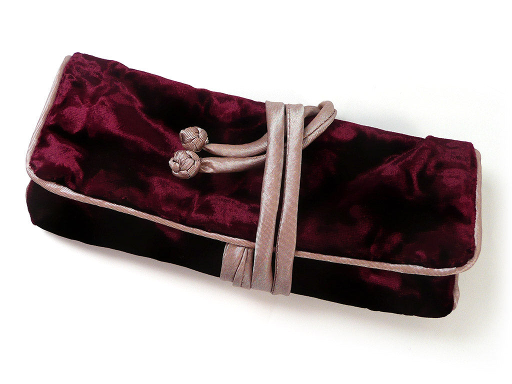 A burgundy velvet jewellery roll with a light beige silky strap closure and decorative knot, shown on a plain white background.