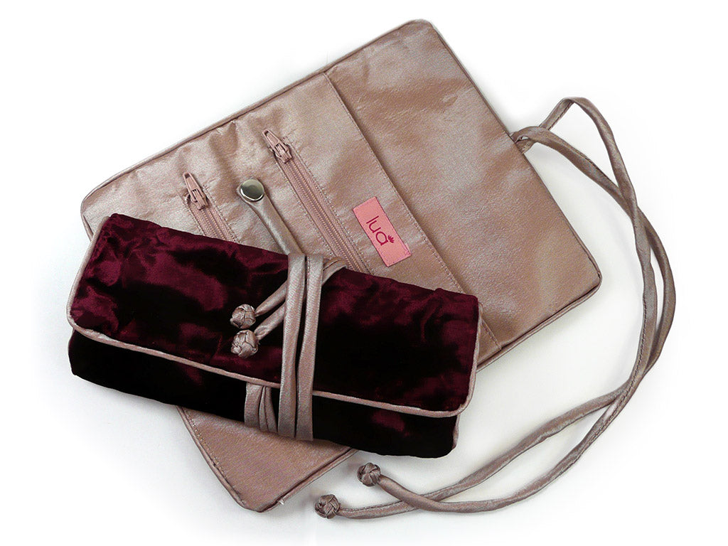 A dusky pink silky lined jewellery roll with a matching tie, shown open to reveal interior compartments and a pink brand tag, with a burgundy velvet pouch tucked inside, on a white background.