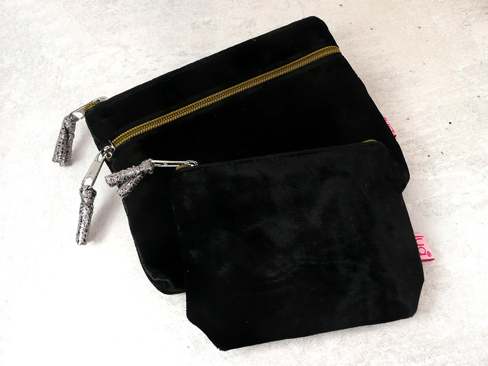 Two black velvet pouches with silver zipper pulls, one larger zippered pouch and one smaller flat pouch, displayed on a light textured surface.
