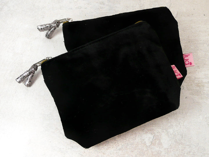 Two black velvet purses with silver zipper pulls and a pink brand tag, displayed on a light textured surface.