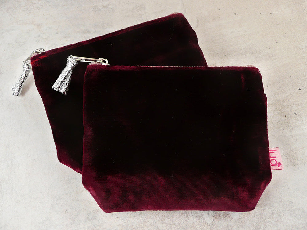 Two deep burgundy velvet zippered pouches with silver zipper pulls, displayed on a light gray textured surface. A small pink tag with the brand name 'Lua' is visible on the front pouch.