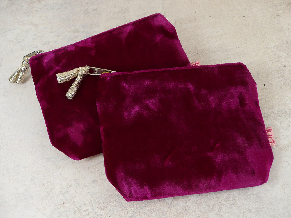 Two small velvet zip pouches in deep cerise fabric, arranged overlapping on a light beige textured surface. One pouch has a silver zipper with a decorative silver tassel pull, and the other has a silver zipper pull with a small pink tag visible on the side.
