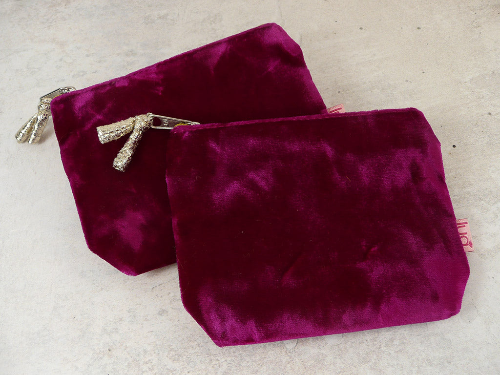 Two small velvet zip pouches in deep cerise fabric, arranged overlapping on a light beige textured surface. One pouch has a silver zipper with a decorative silver tassel pull, and the other has a silver zipper pull with a small pink tag visible on the side.