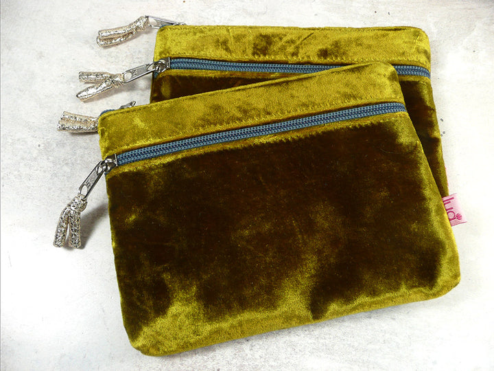Two velvet zip pouches in a mustard yellow-green colour, arranged diagonally on a light grey textured background.
