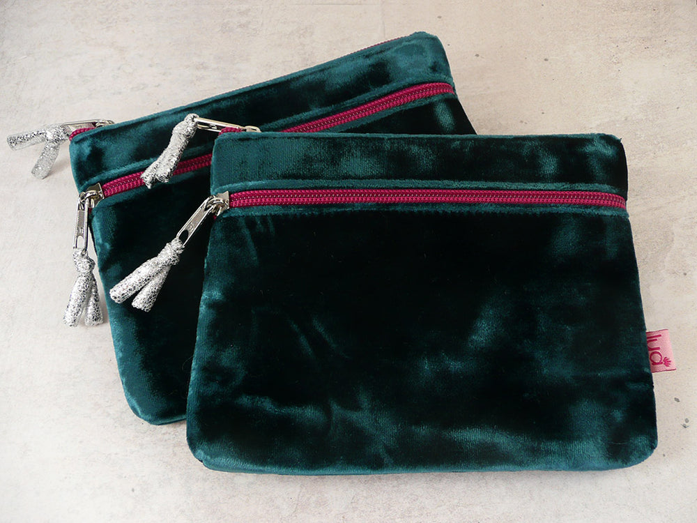 Two dark turquoise velvet zippered pouches with silver zipper pulls and tassel details, arranged on a light beige textured surface.