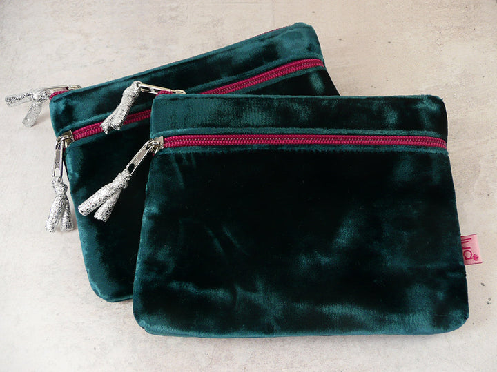 Two dark turquoise velvet zippered pouches with silver zipper pulls and tassel details, arranged on a light beige textured surface.