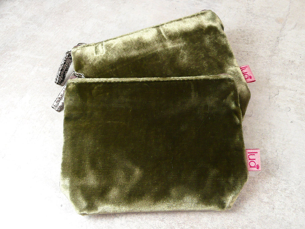 Two olive green velvet zipped pouches stacked on a light gray textured surface, with small pink tags visible on the sides.