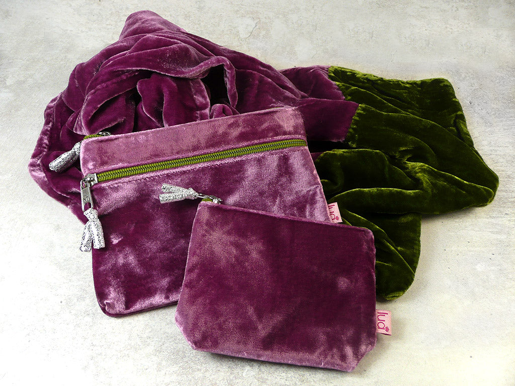 Two velvet zipped pouches and a matching velvet scarf  in plum purple and olive green, arranged on a light grey textured surface.