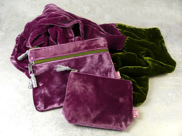 Two velvet zipped pouches and a matching velvet scarf  in plum purple and olive green, arranged on a light grey textured surface.