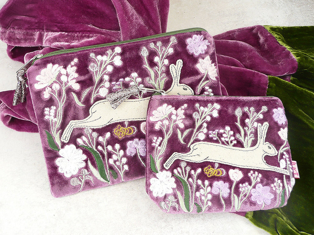 A matching set of purple velvet pouches and a bag featuring embroidered white hares, white and lavender flowers, green leaves, and small gold butterflies, displayed on a light surface with a green velvet fabric prop.