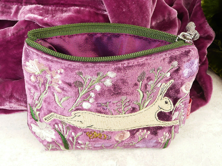 A purple velvet purse with embroidered details of a hare and wildflowers, featuring a green zip, set against a matching purple velvet background.
