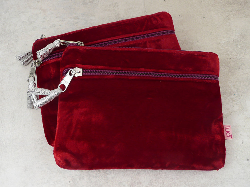 Two red velvet zippered pouches stacked on a light gray textured surface, each with a silver zipper and a decorative silver tassel pull.