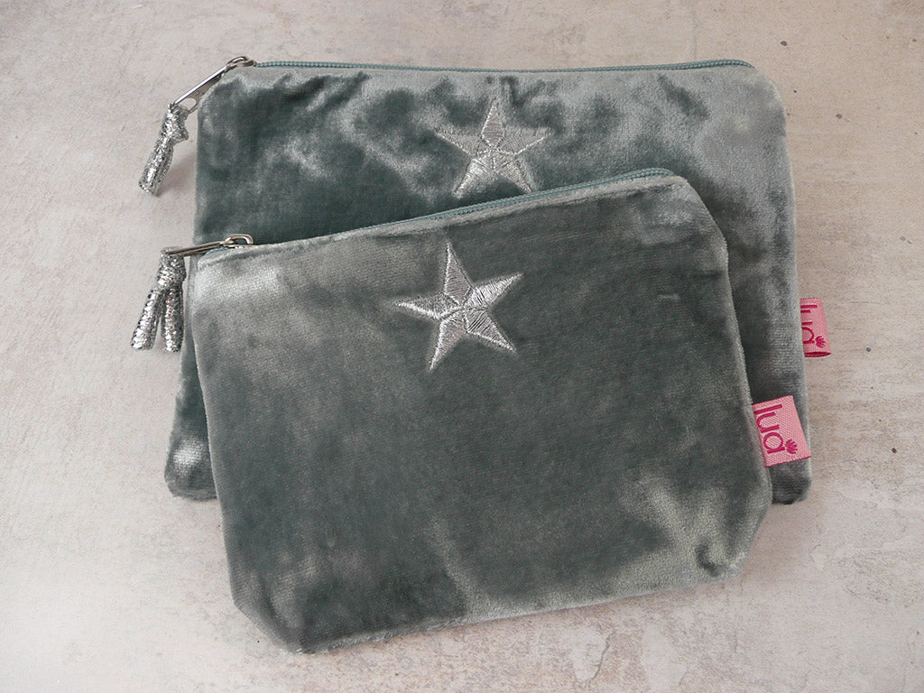 Two blue grey velvet zippered pouches stacked on a light beige textured surface, each featuring a silver embroidered star on the front and a pink fabric brand tag on the side.