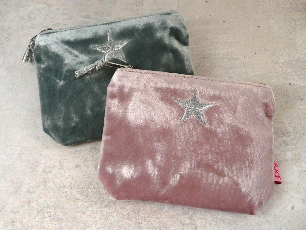 Two velvet zippered pouches on a light stone surface, one in steely blue and one in dusty pink, both featuring a silver star embroidered on the front.