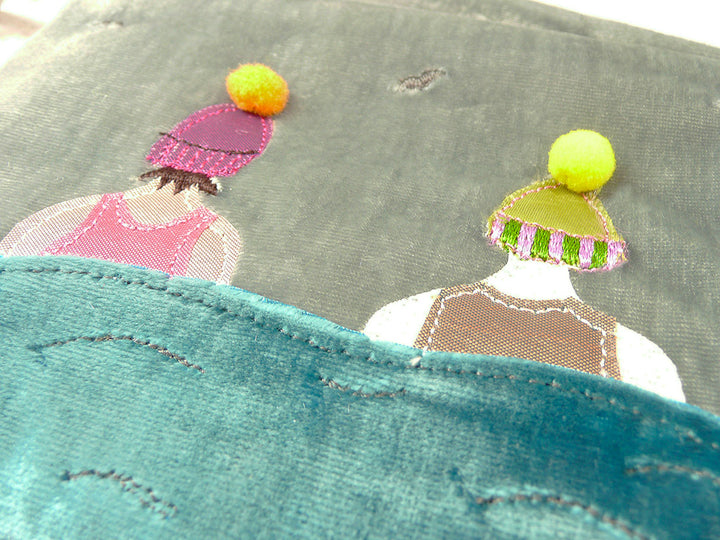 Close-up of a handmade velvet fabric purse, featuring embroidered figures wearing colorful winter hats. The purse shows a teal blue velvet-like front panel with stitched wave details, layered over a grey velvet back with two embroidered characters in pink and brown outfits with yellow and orange pom-poms.