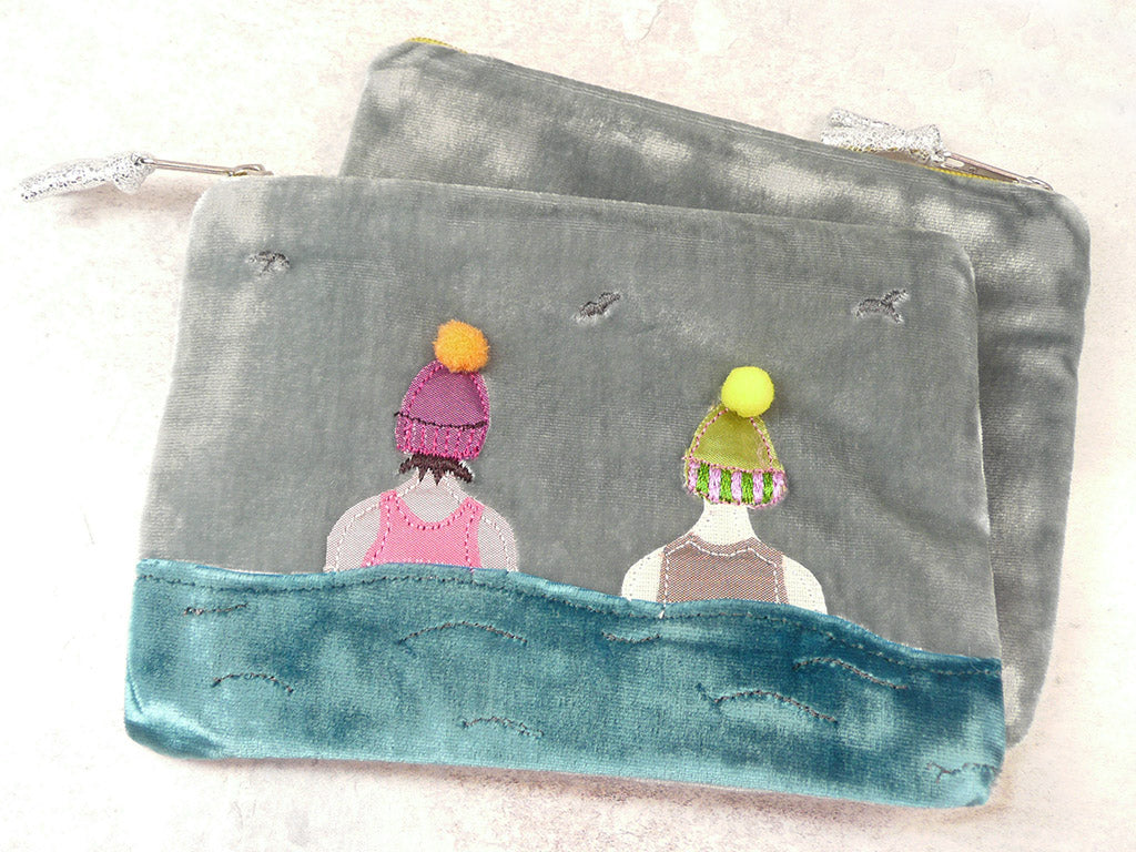 A grey velvet zippered pouch featuring a whimsical embroidered design of two figures seen from behind, wearing colorful knit hats, with a teal blue stitched wave-like border along the bottom.