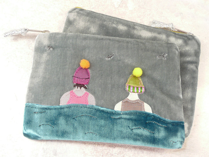 A grey velvet zippered pouch featuring a whimsical embroidered design of two figures seen from behind, wearing colorful knit hats, with a teal blue stitched wave-like border along the bottom.