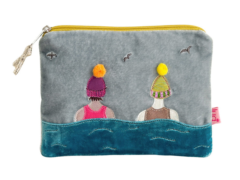 A gray fabric zippered pouch featuring a whimsical embroidered design of two figures in colorful knit hats above stylized blue waves, with a yellow zipper and a small pink fabric tag on the right side.