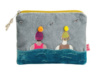 A gray fabric zippered pouch featuring a whimsical embroidered design of two figures in colorful knit hats above stylized blue waves, with a yellow zipper and a small pink fabric tag on the right side.