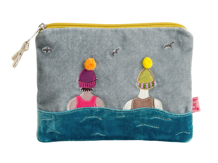 A gray fabric zippered pouch featuring a whimsical embroidered design of two figures in colorful knit hats above stylized blue waves, with a yellow zipper and a small pink fabric tag on the right side.