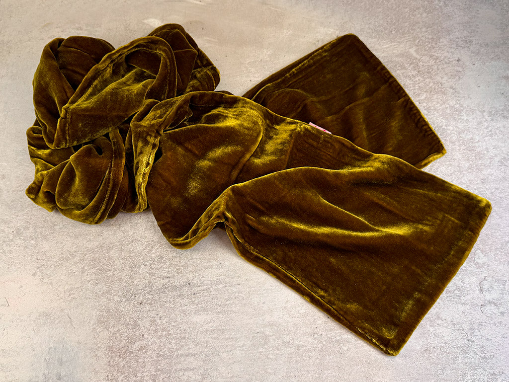 A mustard yellow velvet scarf, styled in a loose knot, shown against a grey concrete background.