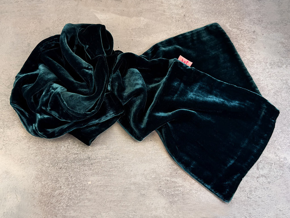A dark teal velvet scarf draped in soft folds on a textured light grey concrete surface.
