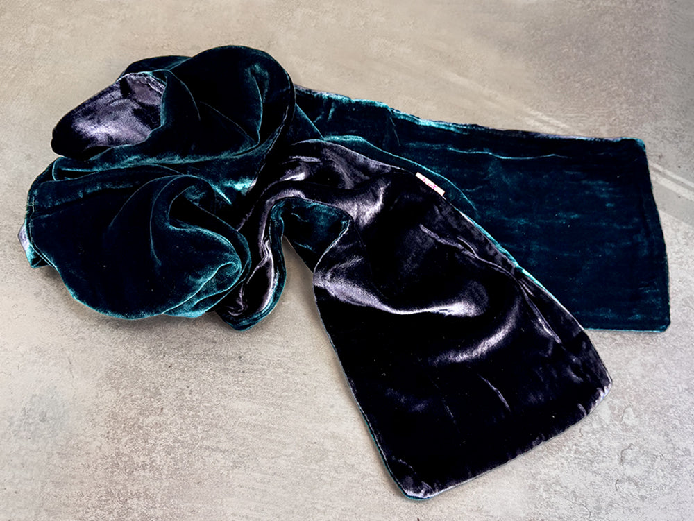 A dark teal velvet scarf with a large bow, laid out on a textured gray concrete surface.