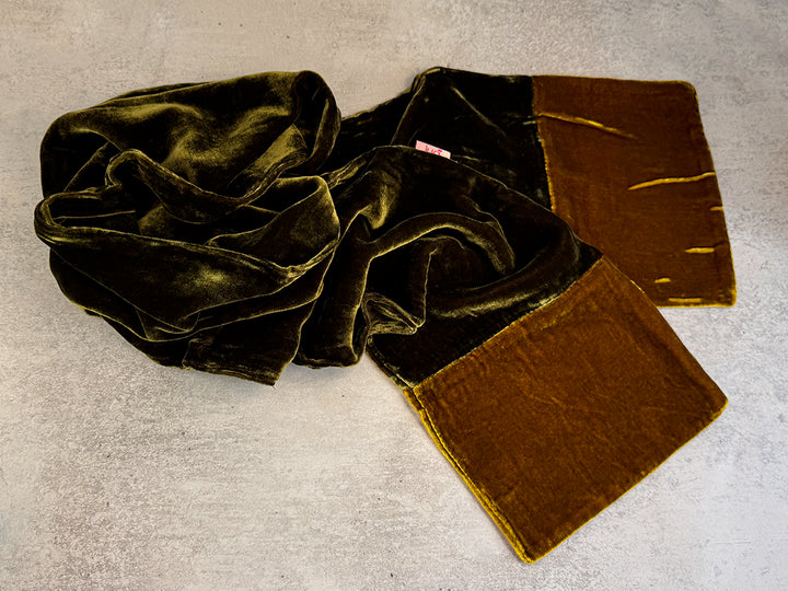A velvet scarf in a two-tone design of dark olive green and mustard green, laid out on a textured light grey surface.
